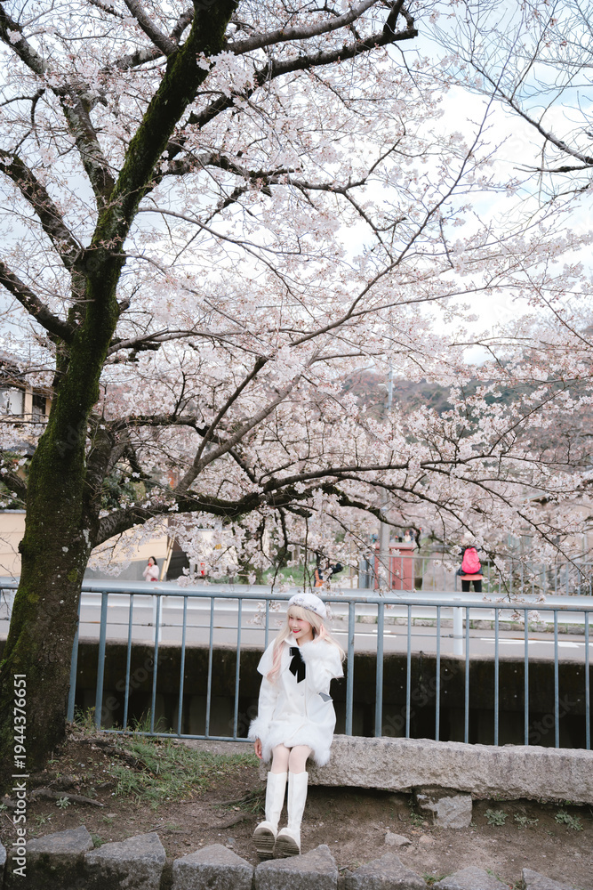 Fototapeta premium Young woman cherry blossom spring tree outdoor sitting white dress boots nature park smiling flower season leisure casual female beauty happiness sunlight peaceful fresh bloom petals trunk grass