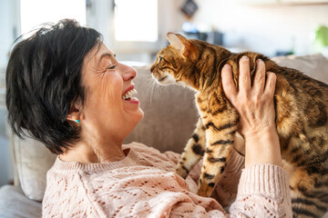 Old woman having fun with her cat sitting on the couch at home - Smiling kitty owner playing in the living room - Animal and people life style concept