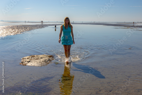 Woman walking on wide tidal flats with beautiful ripple patterns in the sand during low tide. Minimalist coastal landscape with calm sea and clear blue sky.