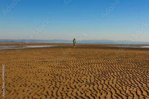 Woman walking on wide tidal flats with beautiful ripple patterns in the sand during low tide. Minimalist coastal landscape with calm sea and clear blue sky.
