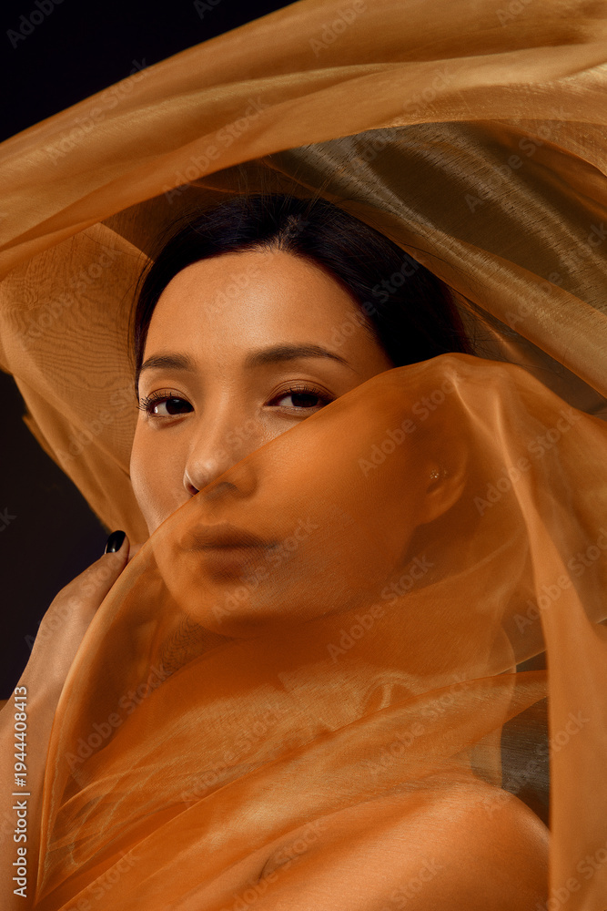 Fototapeta premium Asian woman cinematic portrait with face partially hidden behind translucent golden silk fabric. Luxury aesthetic for cosmetics advertising, beauty branding, fashion editorials and skincare campaigns.