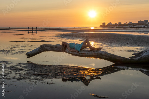 Woman lying on a driftwood log on wide tidal flats with ripple patterns in the sand at low tide. Minimalist coastal landscape with calm sea and setting sun in the background.