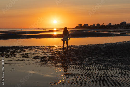Woman standing on a driftwood log on wide tidal flats with ripple patterns in the sand at low tide. Minimalist coastal landscape with calm sea and setting sun in the background.