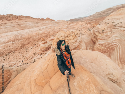 A woman is standing on a rocky hillside, taking a selfie with her cell phone