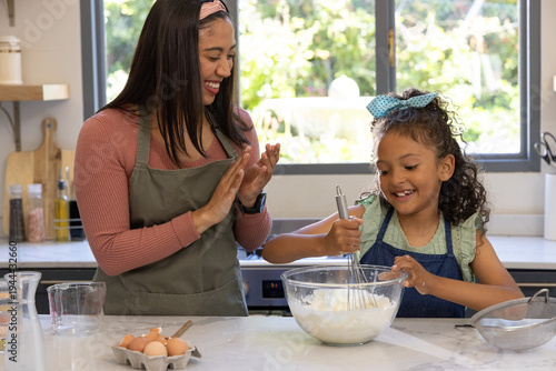 Mother and child baking at marble counter in kitchen whisking batter in glass bowl wearing aprons