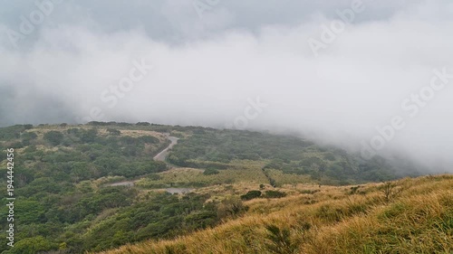A scenic view of a winding road through the grassy hills of Datun Mountain in Taipei, Taiwan, surrounded by moving clouds and mist at twilight.