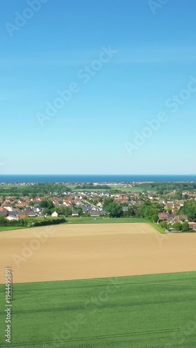 Wide view of the Normandy countryside with cultivated fields, a village, and the distant coastline under a clear blue sky. Rural landscape in northern France.