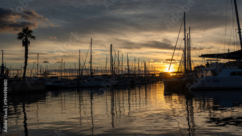 Dramatic sunset over Alicante marina with sailboats silhouetted against colorful sky, calm reflective water, and peaceful harbor atmosphere for travel, tourism, and coastal lifestyle concepts.
