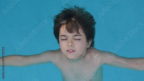 Happy Child relaxing in blue swimming pool. Boy with dark hair floats on his back in clear, bright blue water. His eyes are closed in peaceful relaxation. Serene, calm mood. Tranquility and summer.