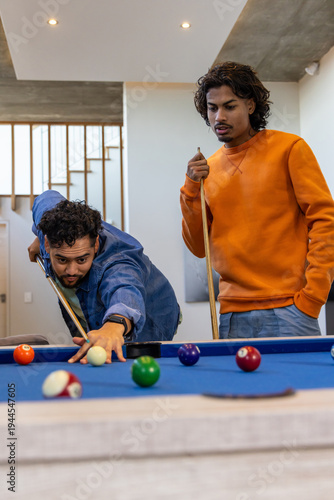 Indian friends playing pool in home rec room, leaning and lining up shot on blue-felt table