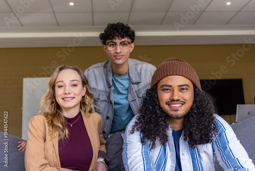 Diverse coworkers smiling and sitting on sofa in lounge, showing laptop, whiteboard, monitor