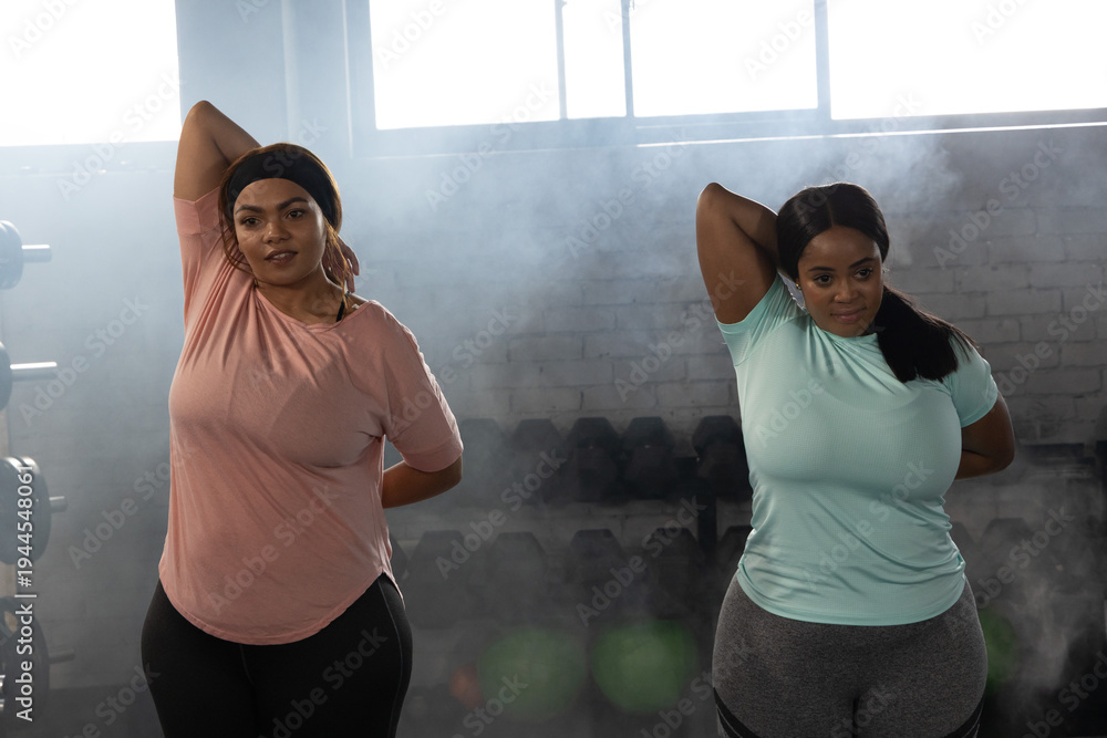 Naklejka premium African American female friends stretching in gym beside dumbbell rack, wearing pink and blue tops