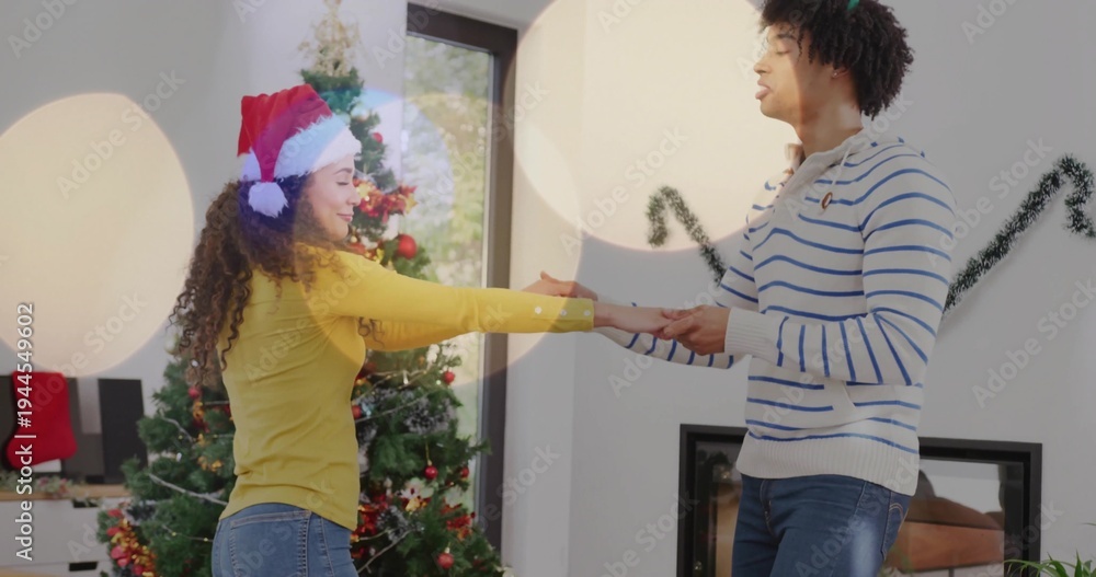 Naklejka premium Dancing couple holding hands in living room, wearing red Santa hat and sweaters near Christmas tree