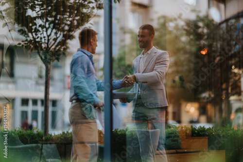 Принти на полотні Two professionals shake hands outside a modern office, seen through a glass storefront in the city