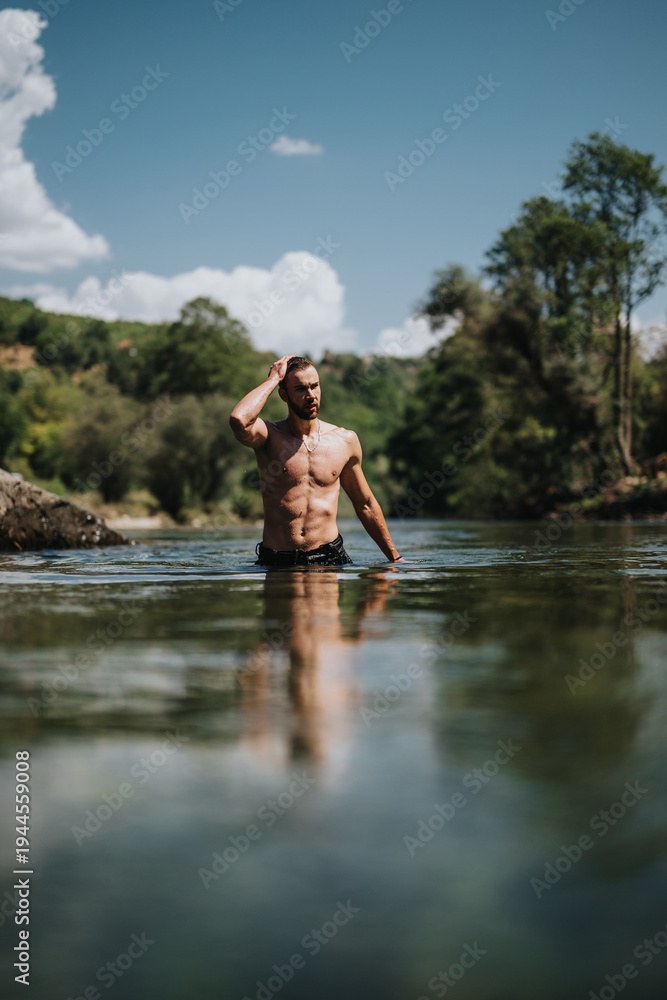 Fototapeta premium A fit man stands in a calm river, adjusting his hair. The sunny outdoor scene features lush trees, clear water, and a bright blue sky.