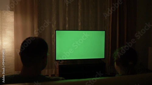 Family watching television with blank green chroma key screen in dark living room. People watch TV with green chroma key screen. Family Sitting Together Sofa In Their Living Room Night Watching TV.