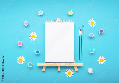 White canva with paintbrushes, paints and fresh spring flowers on blue background. Creativity, artistic inspiration and celebration of World Art Day