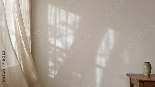Sheer curtains blowing in the wind by a window with sunlight shadows falling on a light wall and a wooden table.