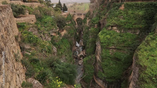 A cinematic high angle shot looking down into the lush, green El Tajo canyon. The footage shows the river flowing between vertical cliffs with the ancient Puente Viejo stone bridge as a focal point.