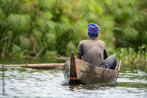 Local fisherman paddling a wooden canoe on a calm wetland lake (lake Victoria) near Entebbe, Uganda. Traditional fishing scene surrounded by lush green vegetation in East African nature.