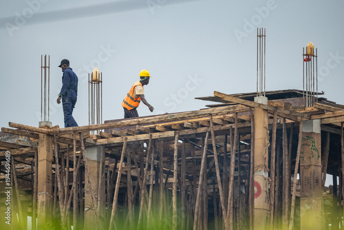 Construction workers wearing safety helmets working on a building site near Entebbe, Uganda. African builders installing wooden formwork on a structure under clear blue sky.