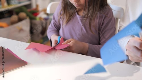 Young child focusing on cutting folded pink paper with blue handle scissors at a white table, engaging in a school project or home craft activity with concentration and developing fine motor skills