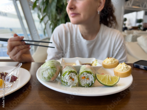 Woman using chopsticks to eat delicious fresh spring rolls with lime in a modern airport lounge, relaxing before travel