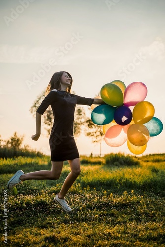 Joyful young woman jumping with colorful balloons in sunset field