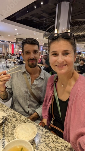 Multiracial couple with arabic man and caucasian woman taking selfie together at table, friends group laughing and enjoying chinese food lunch in indoor eatery, bonding and happiness moment