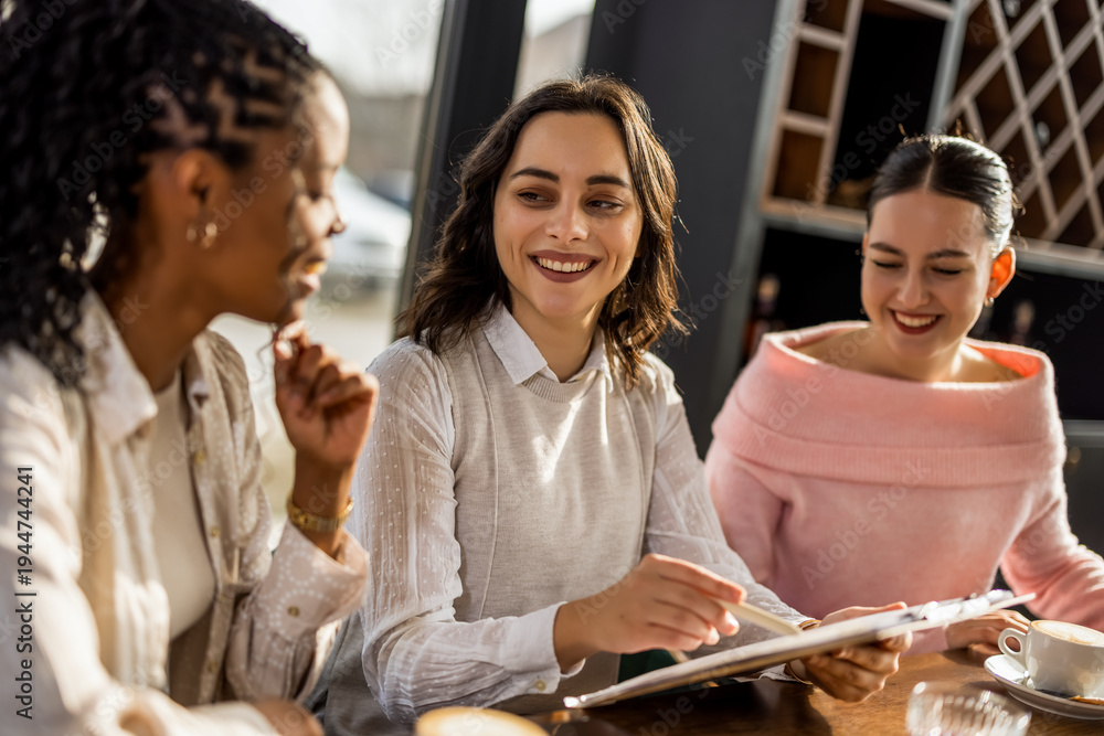 Fototapeta premium Three cheerful diverse business women laughing while reviewing project documents in a cafe.