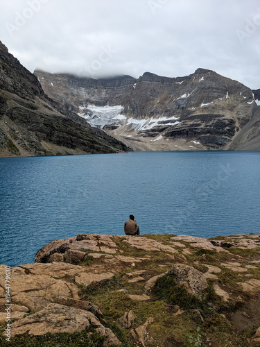 A man sits on the edge of a cliff admiring Lake McArthur and the Rocky Mountains. Glaciers and snow rest on the mountains during a cloudy day in the summer.