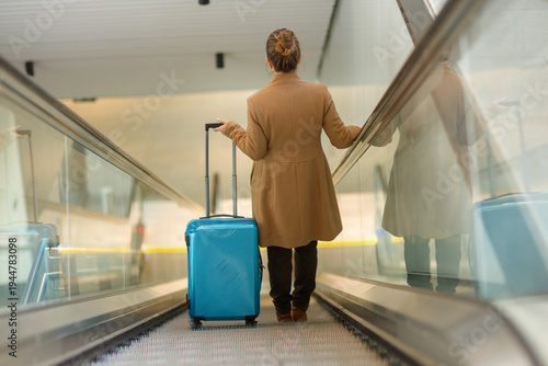 A woman in a tan coat pulls a blue rolling suitcase while moving up an escalator in a bright, modern transit station during her journey.