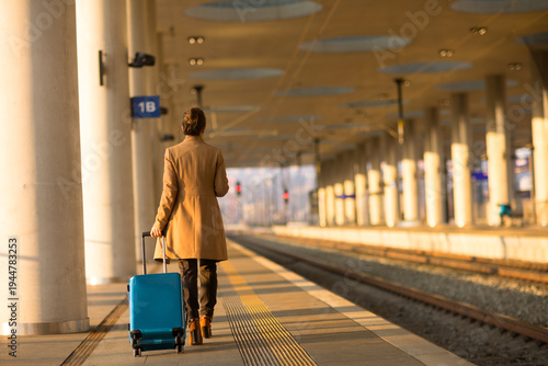A person in a tan overcoat walks along a railway platform with a blue rolling suitcase, heading toward their departure gate in the warm glow of the sun.