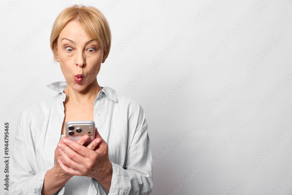 Fototapeta premium Surprised mature woman with short blond hair looks at smartphone with wide eyes and pursed lips. Isolated on white background. Concept of shock, message, and technology use.