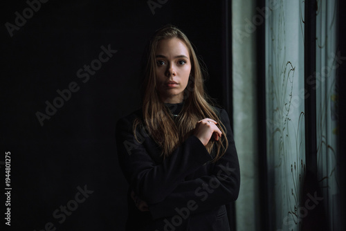 Low key portrait of a mysterious young woman in a black jacket posing near a window in the dark