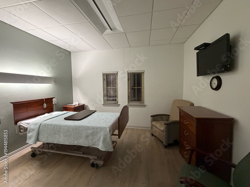 A private patient room in a skilled nursing or rehabilitation facility, shown from a wide angle. Features a hospital-style adjustable bed with a decorative headboard, overbed tray table, wall-mounted 