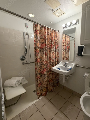 A functional accessible bathroom in a senior care or skilled nursing facility, featuring a roll-in shower with grab bars, handheld showerhead, shower chair with folded white towels, pedestal sink with