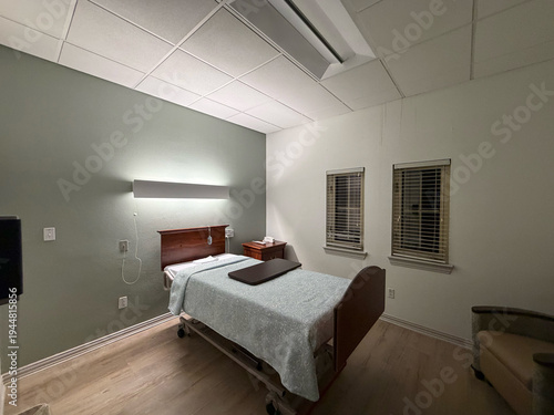 A close-up view of the bed area in a private skilled nursing or rehabilitation room. Adjustable hospital bed with decorative wood headboard, soft blue patterned bedding, overbed tray table, wall-mount