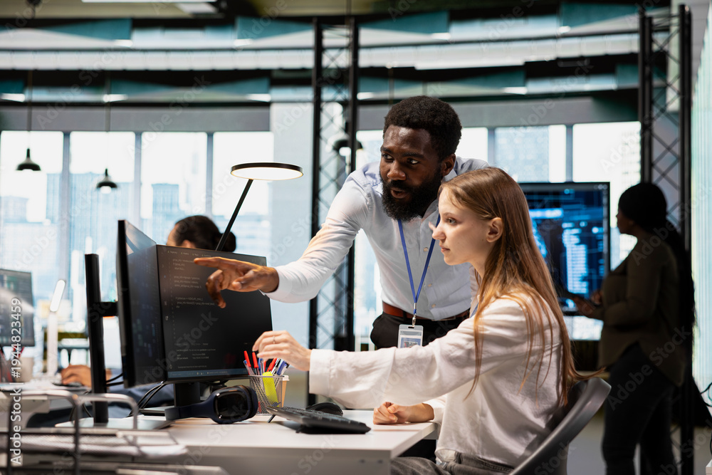 Fototapeta premium Lead programmer providing support to new worker using PC in software development agency. African american IT expert helping trainee coding during internship in tech startup