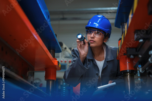 Indian female industrial engineer uses flashlight to inspect machinery in a manufacturing factory for quality control purposes.