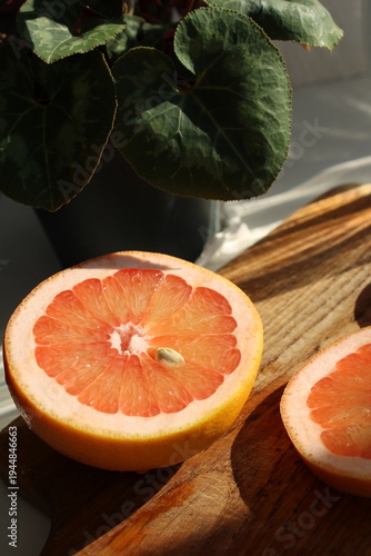 Fotografie Fresh pink grapefruit halves on a rustic wooden cutting board with warm sunlight and shadows, featuring green plant leaves in the background