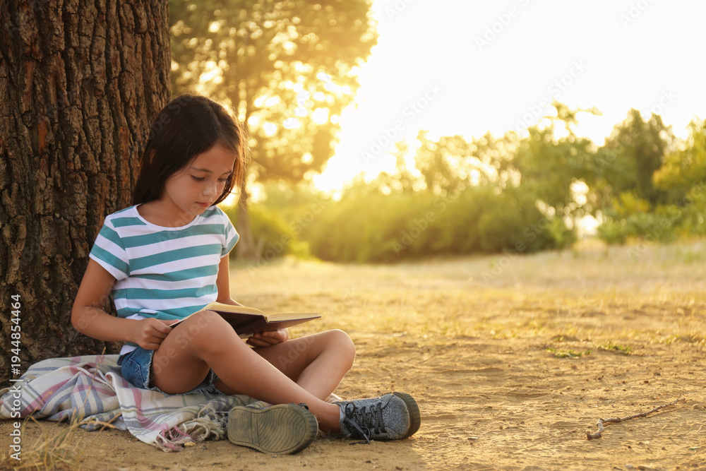 Naklejka premium Cute little girl reading book near tree in park