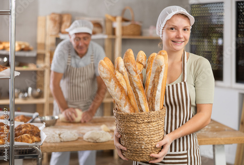 Slika na platnu Smiling young female baker holding basket of golden crispy baguettes, proudly pr