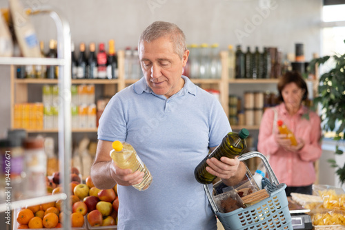 Photography Elderly man buyer chooses olive or sunflower oil in bottle in grocery store