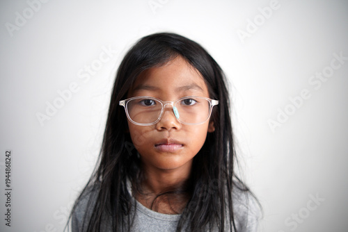 Little Asian girl with eyeglasses looking at camera with neutral expression, standing indoors, blank stare, serious childhood face