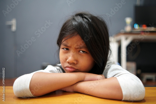 Moody Asian little girl sulking and leaning face on table, looking at camera with grumpy expression, unhappy child feeling bored or disappointed