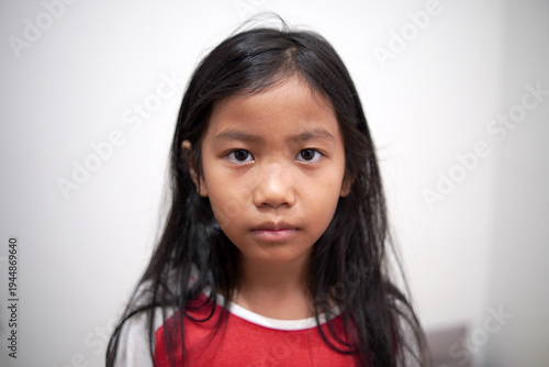 Little Asian girl looking at camera with neutral expression, standing indoors, blank stare, serious childhood face
