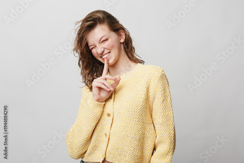 Fotografie Young woman in yellow knitted sweater smiles and holds finger to lips in a shushing gesture