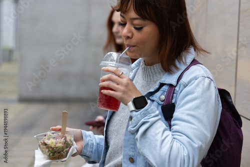 Two women standing by tiled wall on sidewalk sipping red drink holding salad bowl