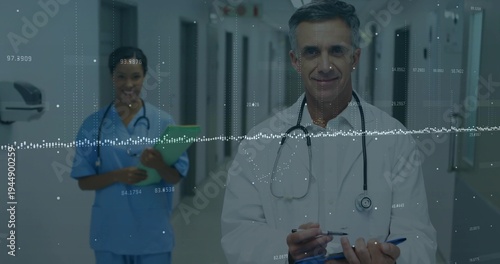 Standing senior doctor with stethoscope in labcoat in hospital hall, blue-scrub nurse, data overlay